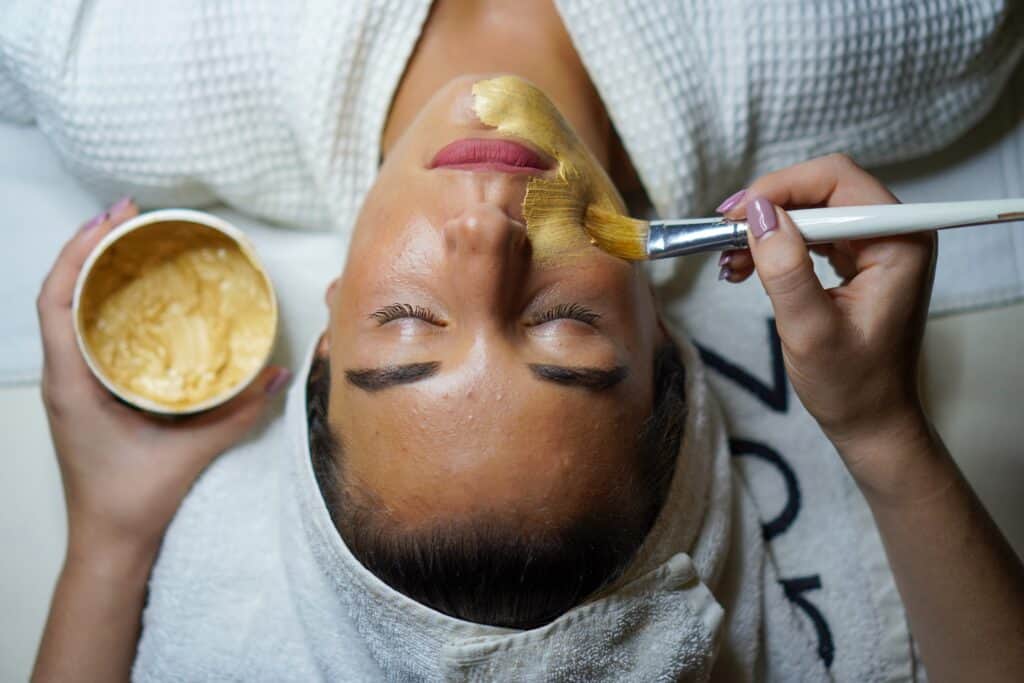 A woman lies down with eyes closed as someone applies a face mask on her