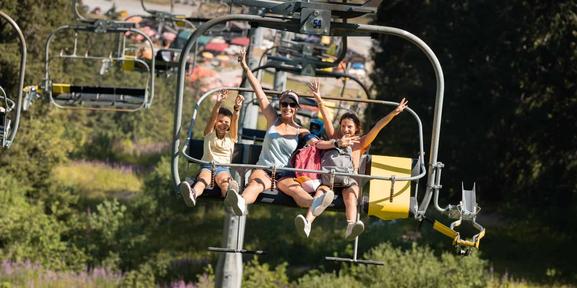 Une mère et ses deux filles lèvent les bras sur un télésiège l'été en montagne.