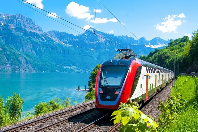 A train runs along the edge of a lake with mountains in the background and a blue sky