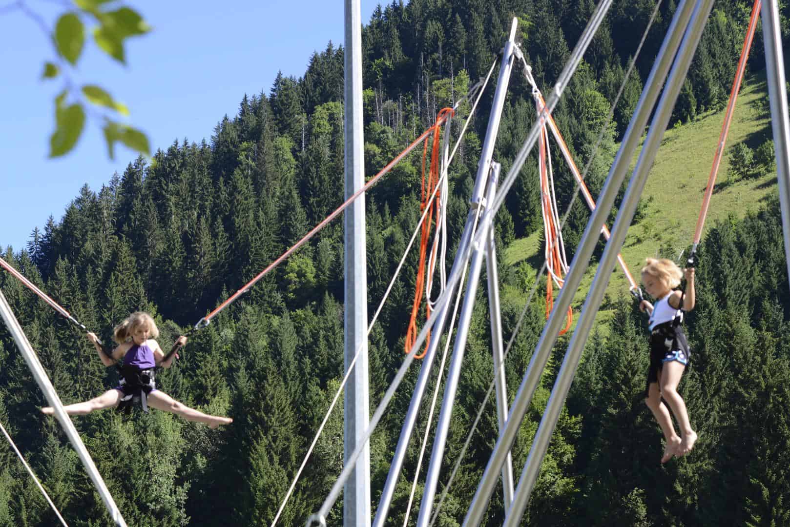 Two little girls try out the bungee trampoline in front of a mountain slope