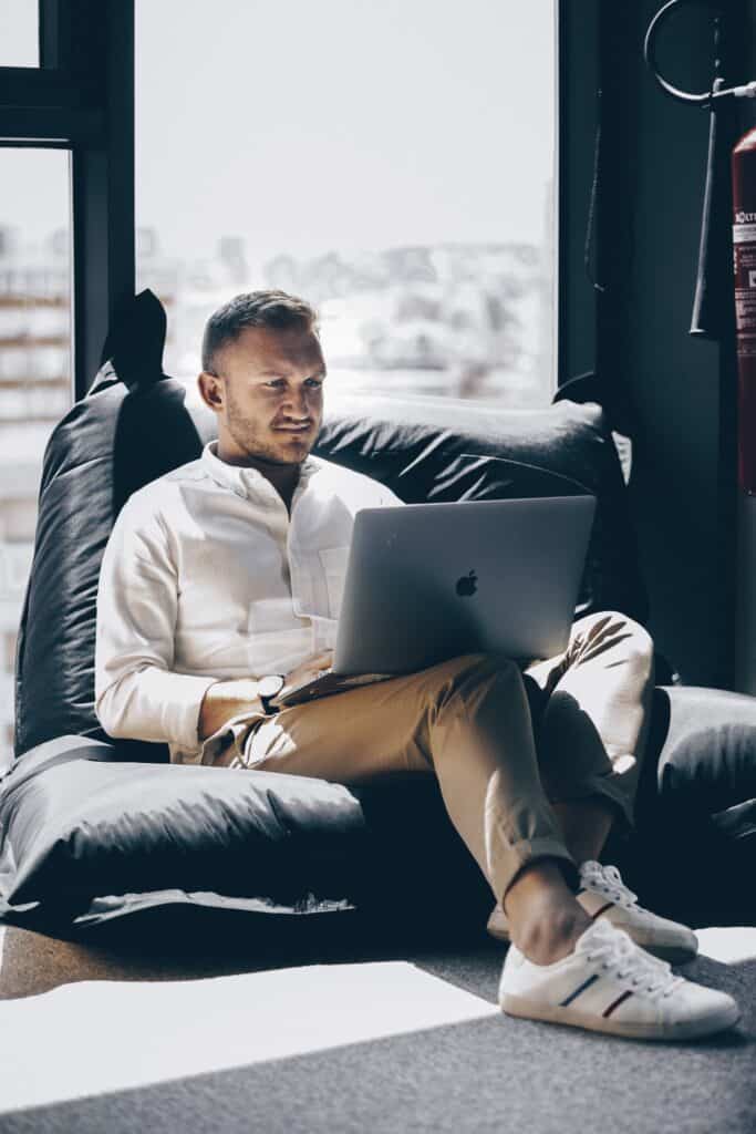 Un homme assis sur un fauteuil gris travaille avec son ordinateur portable sur les genoux.
