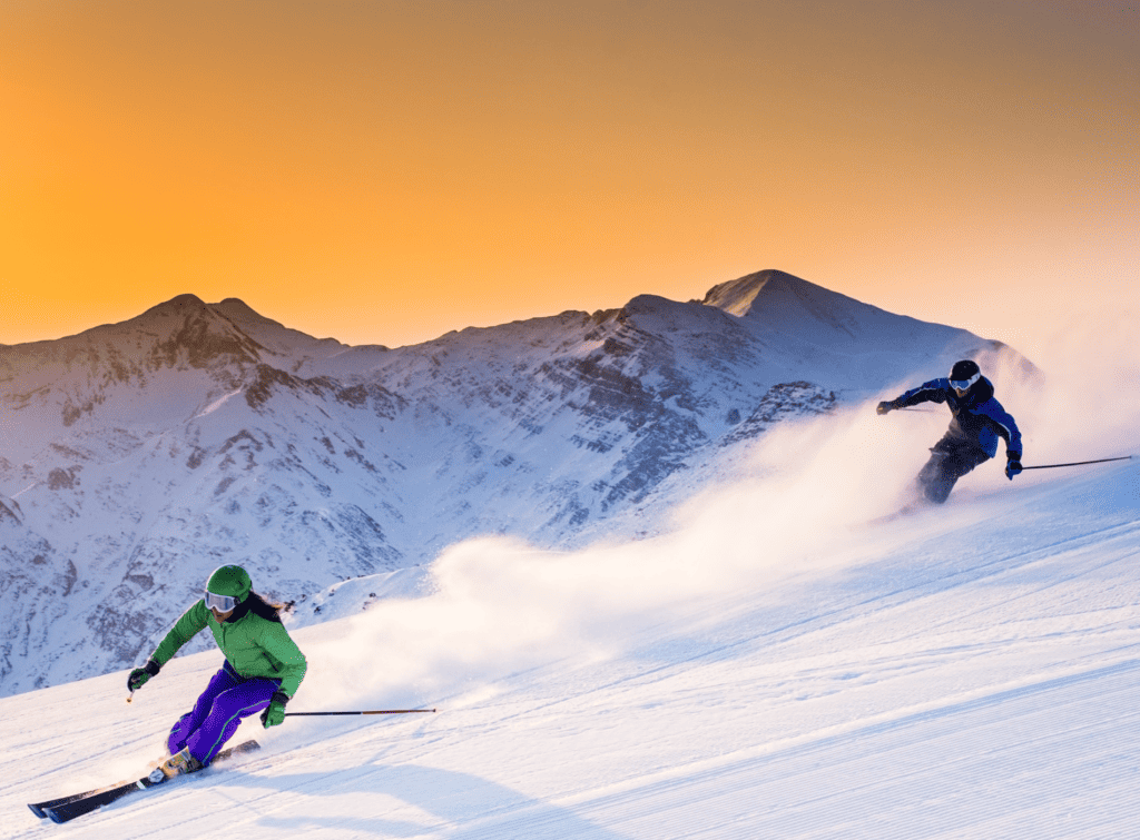 Two people ski down a slope at sunset