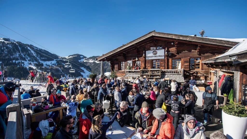 Une foule célèbre l'après-ski sur la terrasse ensoleillée du Chalet des Praz, avec les pistes en arrière-plan.