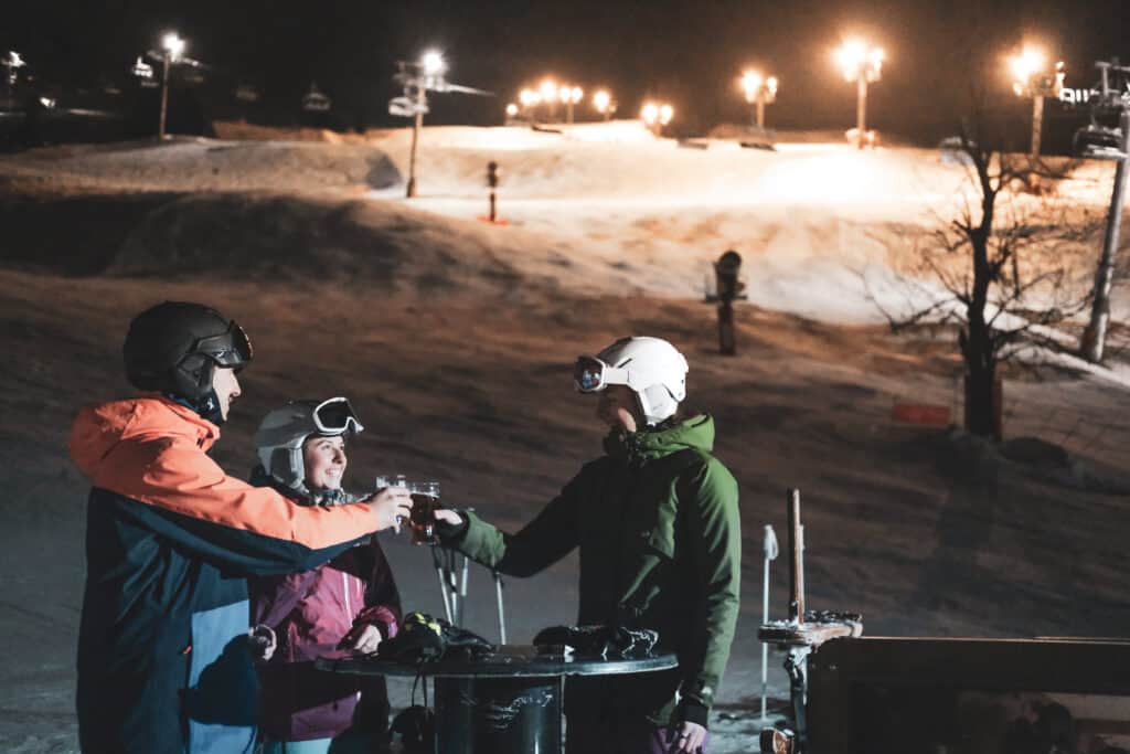 Three people drink vin chaud by the piste at night
