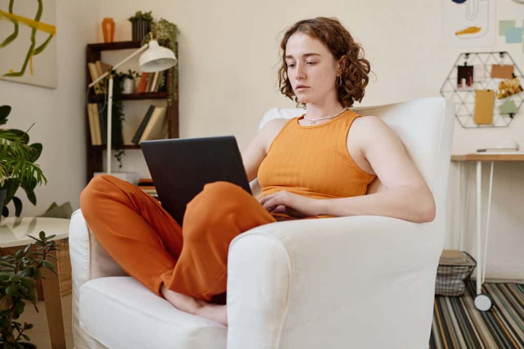 Une femme portant un haut orange assis dans un fauteuil avec son PC portable. 