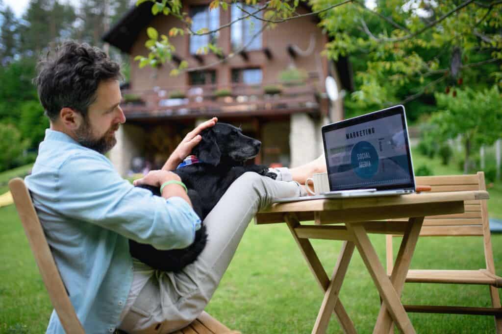 Un homme assis dans le jardin d'un chalet utilisant son ordinateur avec son chien assis sur ses genoux. 