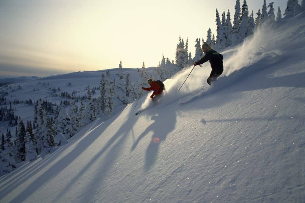 Two people ski down a ski slope as the sun sets