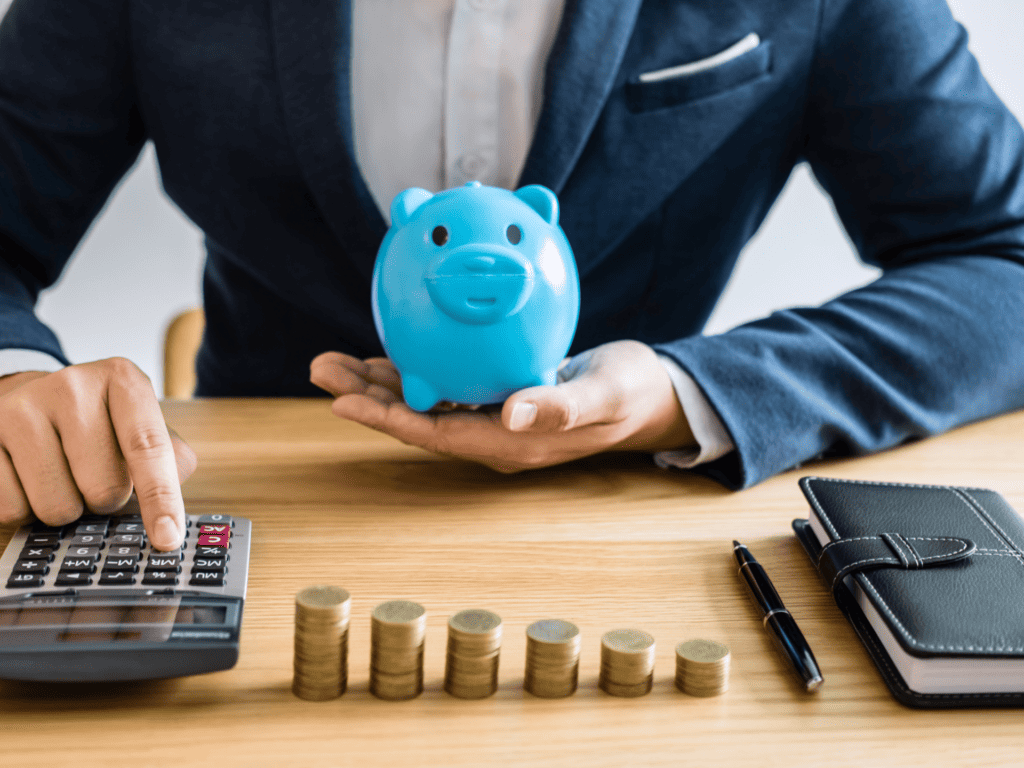 A man in a suite tapping on a calculator while holding a blue piggy bank