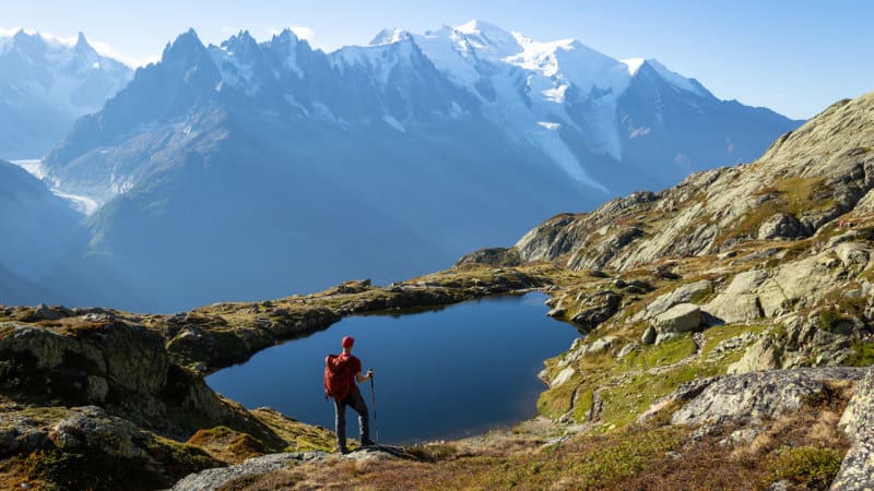 A walker stands next to a mountain lake and contemplates a view of the mountains