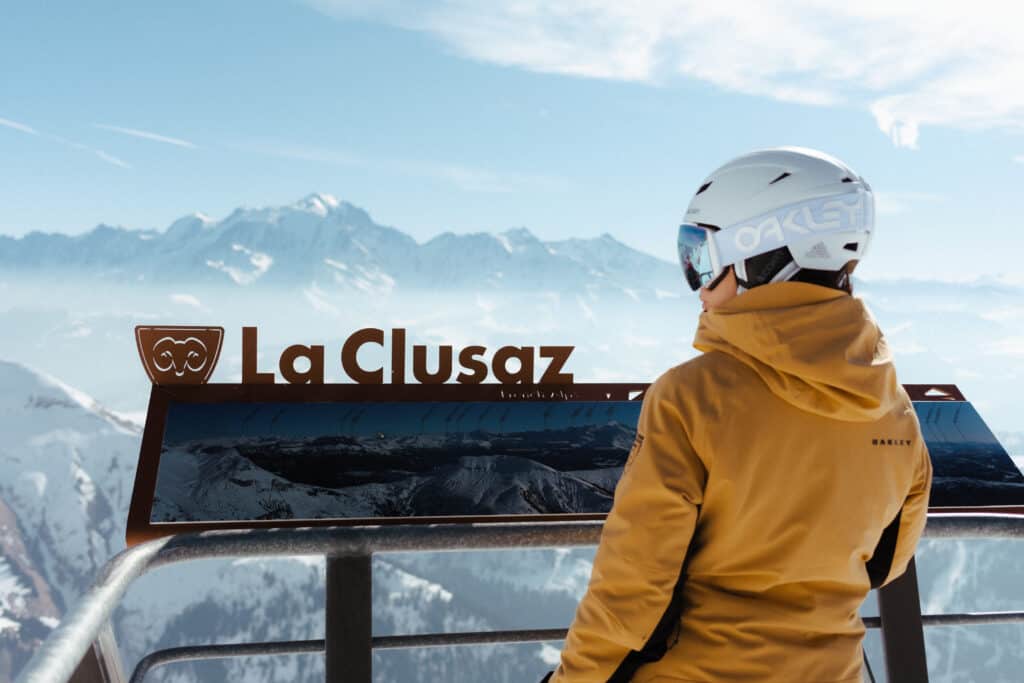 A person wearing a yellow ski jacket stands in front of a La Clusaz map in the mountains