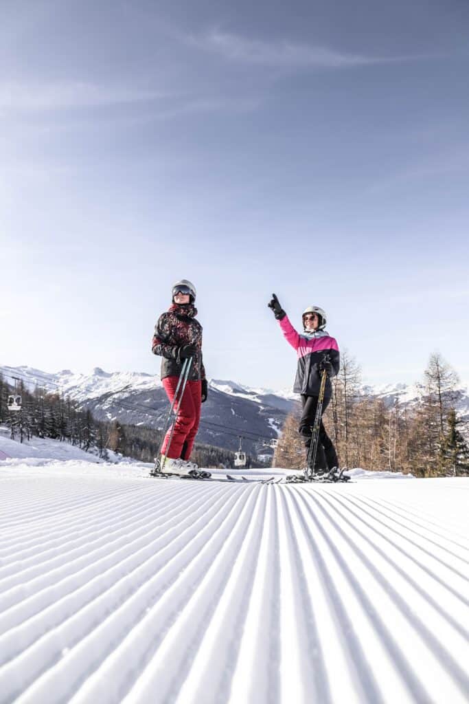 Two people on skis stand on a groomed piste