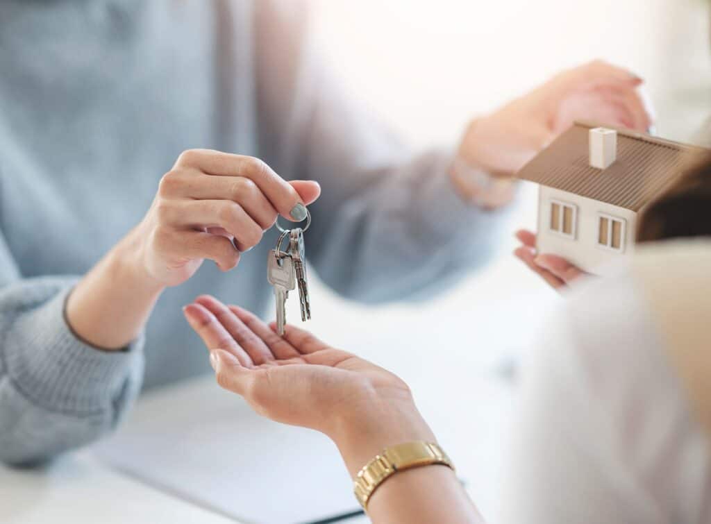 Mains de personnes échangeant des clés et une maisonnette en bois.