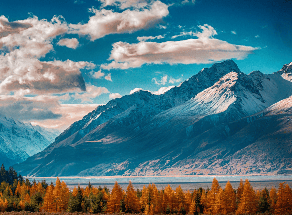 A view across a lake to snowy mountains, clouds in the sky and trees in autumn colours in the foreground