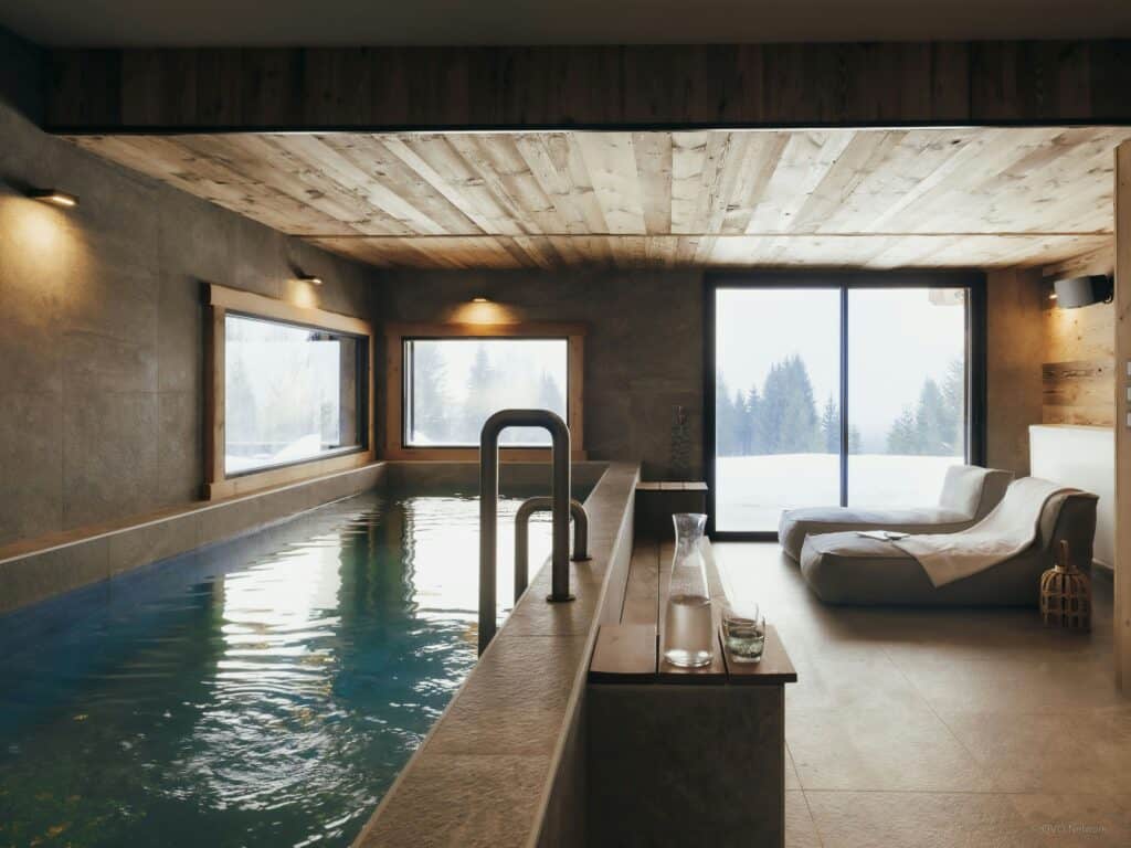 Indoor swimming pool and resting chairs surrounded by bay windows and a snowy landscape.